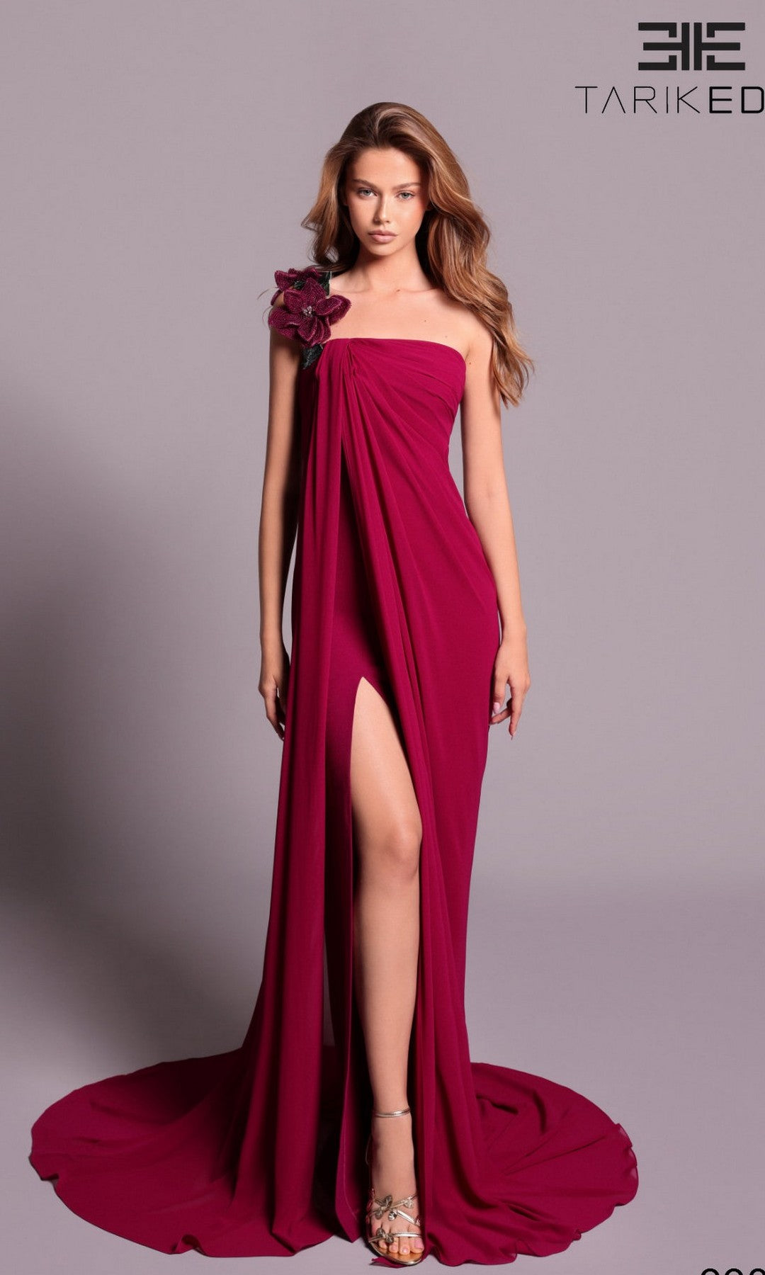 Woman wearing the Tarik Ediz 99036 long formal dress in sangria, strapless with a high slit, floor-length draping, and 3D flower on one shoulder, posed against a plain background. TARIK ED logo appears in the top right corner.